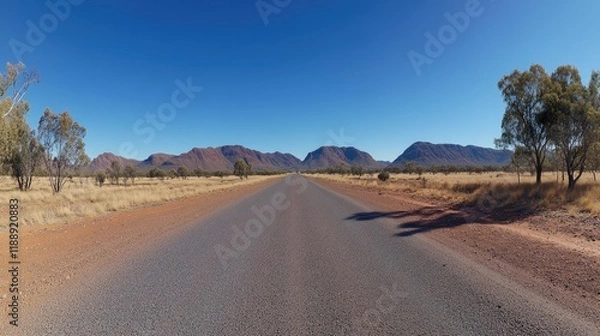 Fototapeta A wide-angle photo of the empty road in Australia's Outback, with red earth and sparse vegetation on both sides, a clear blue sky, and distant hills in the background.