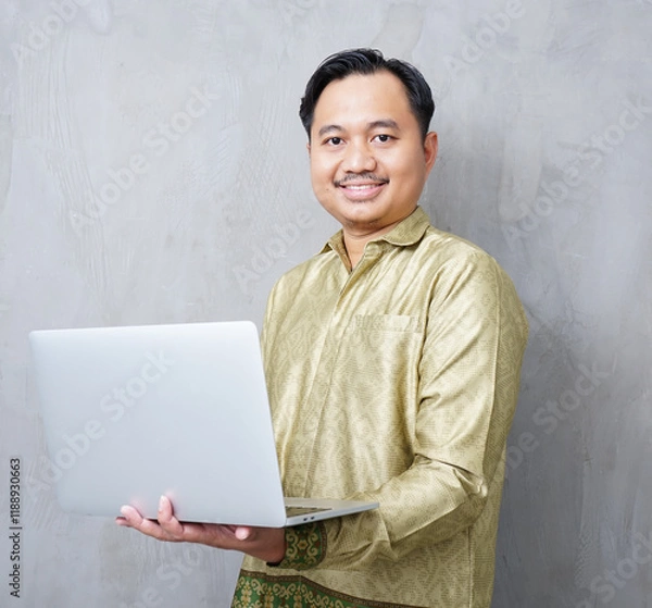 Obraz Adult Asian man wearing batik smiling at camera while using his laptop - Indonesia men