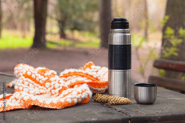 Obraz thermos and scarf on wooden table with nature on background. Tou