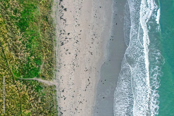 Fototapeta A serene top-down view of an Irish beach, showcasing gentle waves meeting the sandy shore and lush coastal grasslands