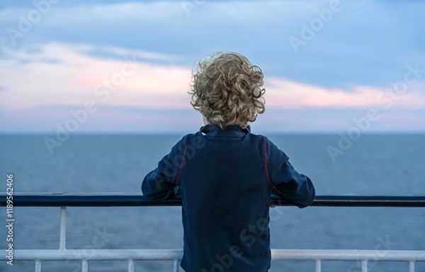Fototapeta A young child with curly hair stands on a ferry, resting his arms on the railing, gazing at the distant horizon. The sky displays soft hues as the sun sets over the water