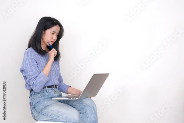 Fototapeta A young woman is working on her laptop, holding a pencil in one hand, with a thoughtful expression on her face