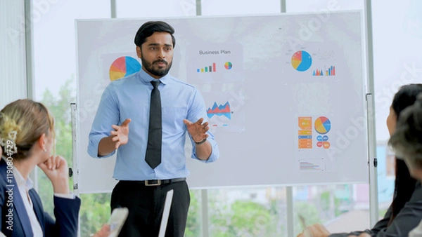 Obraz Young indian business manager standing and presenting to multiracial group of diversity colleagues in front of white board in meeting at boardroom in office . leader