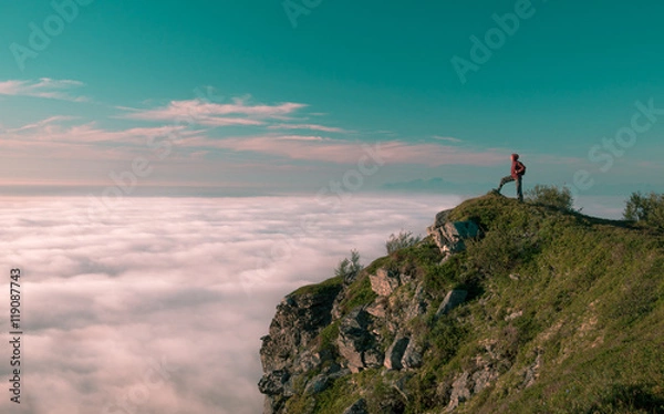 Fototapeta Toned image adult woman with a backpack stands on the edge of a cliff and looking at the sunrise against the blue sky and thick clouds floating down