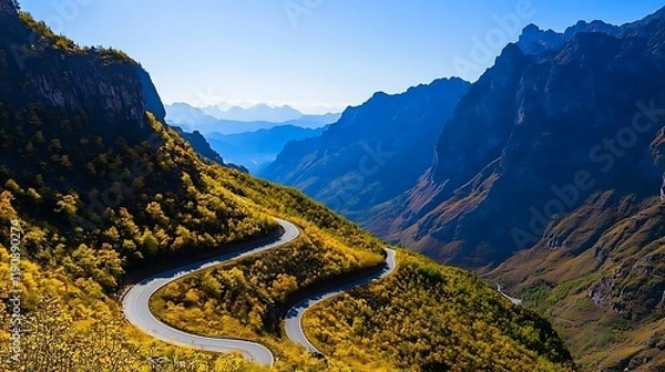 Fototapeta Scenic winding road through vibrant autumn mountains with distant peaks and clear sky