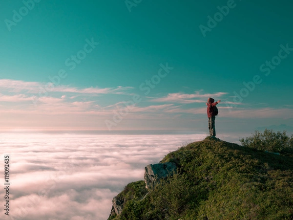 Fototapeta Toned image adult woman with a backpack stands on the edge of a cliff and looking at the sunrise against the blue sky and thick clouds floating down
