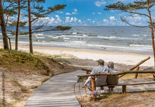Obraz Happy couple of seniors are resting in pine forest and looking at a distance near a sandy beach of the Baltic Sea. Photo depicts ecological tourism in Baltic region. No AI tools were used