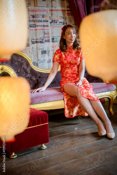 Fototapeta A woman in a red dress sits on a couch in front of a wall covered in Chinese newspapers