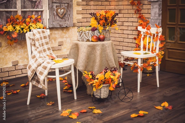 Fototapeta A cozy patio. Autumn leaves lying on the wooden floor, at the center are two white chairs and a table with burlap, fruits and a vase of berries and foliage. Background is brick wall and vintage window