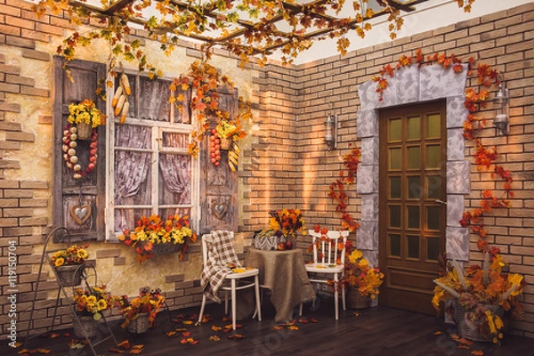 Fototapeta Patio. Shutters of the window and brick walls decorated with autumn garlic, pepper, corns and yellow leaves. There are two chairs, table with fruits in frame.