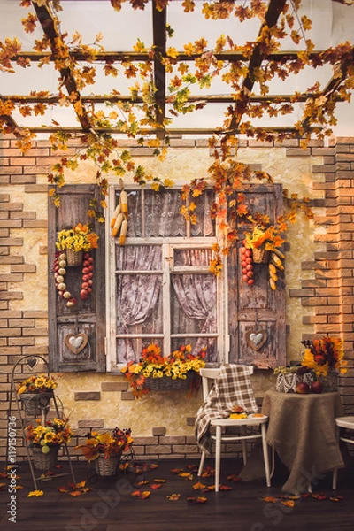 Fototapeta Evening patio. Window with vintage shutters decorated with autumn vegetables. Yellow leaves everywhere, at the center two chairs and a table with burlap, fruits and a vase of berries and foliage.