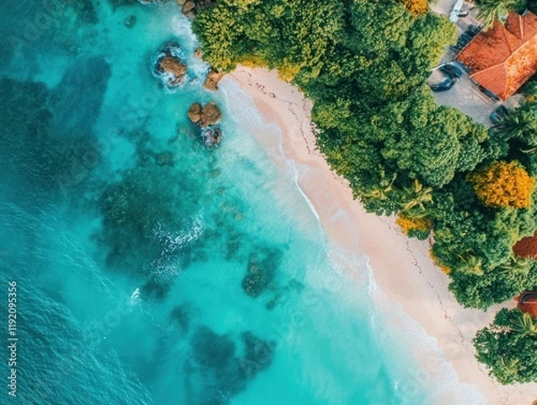Fototapeta Aerial view of a turquoise ocean meeting a white sandy beach with green trees and red roofed buildings