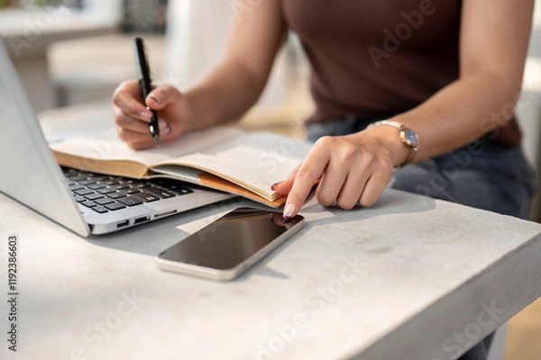 Fototapeta A close-up hand of a woman using her phone while taking notes in a book and working on her laptop.