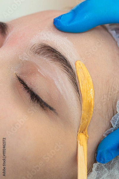 Fototapeta Hands of Professional Beautitian While Completing Eyebrow Correction with Wax and Wooden Sticks in Beauty Salon As Eyebrow Hair Removal with Wax.