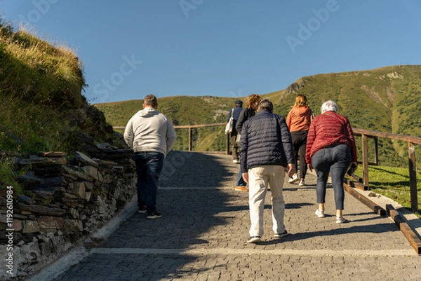 Fototapeta A group of people are walking up a hill. The hill is covered in grass and rocks. The people are walking on a stone path. The sky is blue and clear