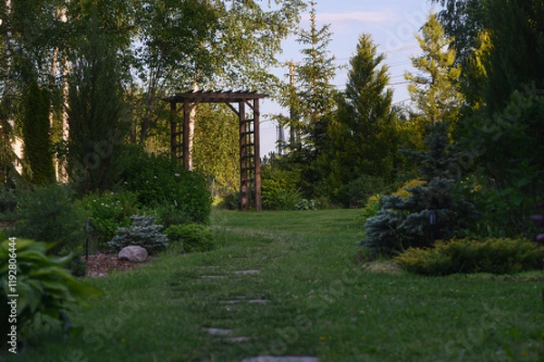 Obraz Beautiful summer garden view with curvy stone pathway and wooden archway (pergola). Natural rustic cottage garden with hostas, conifers and shrubs
