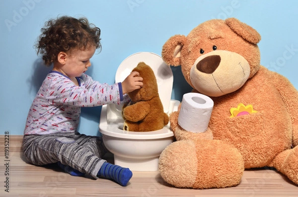 Fototapeta Dziecko siedzące na podłodze ucząc małego misia, który siedzi na nocniku. Cute Kid Potty Training dla siusiu i kupy pomocy przez miś, który daje mu papier toaletowy