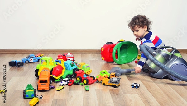 Fototapeta Mother cleaning the messy room full of toys. Close up on woman's hands cleaning with a vacuum cleaner. Upset kid playing with many toys on the floor