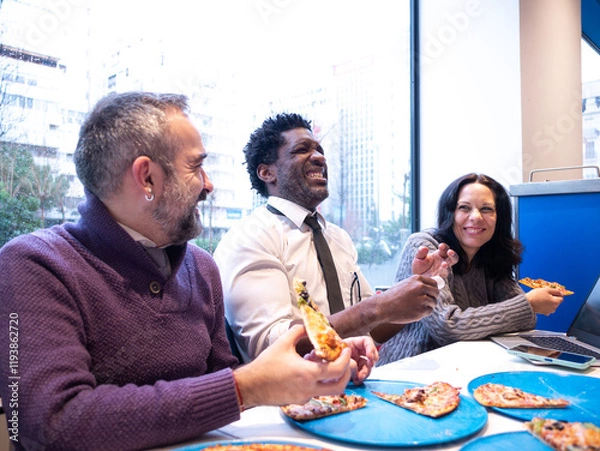 Fototapeta Three colleagues laugh heartily while enjoying pizza around a table with a laptop and phone, set against a bright city backdrop, embodying teamwork and a positive workplace vibe