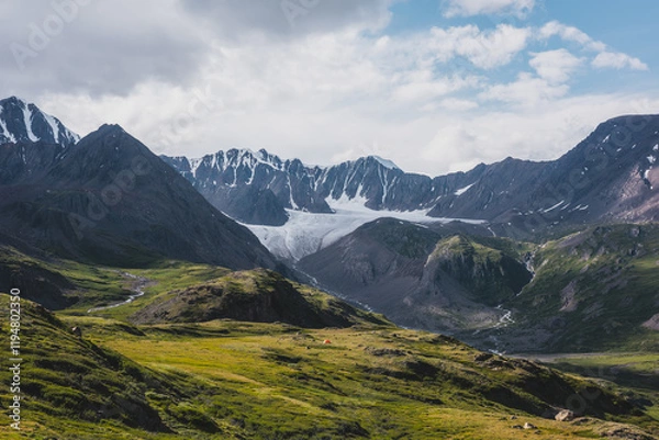 Fototapeta Dramatic scenery in alpine valley with creek among green hills and rocks with view to rocky pointy peak, large snow-capped peaked top, mountain range and big glacier tongue under clouds in blue sky.