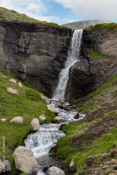 Fototapeta Waterfall on the mountain in black stone that forms a river in a green valley with stones
