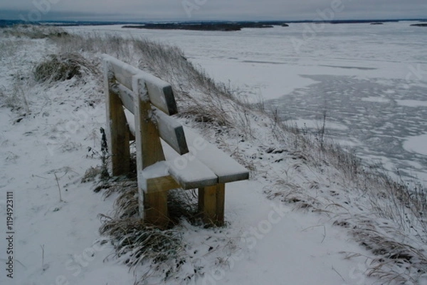 Fototapeta bench in the snow on a high bank near a frozen river