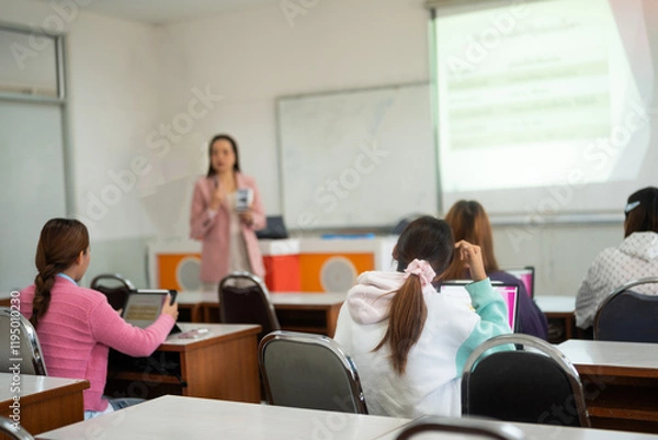 Fototapeta Students engage in a lecture, sitting at desks with their attention focused on the lecturer at the front of the classroom. The lecturer uses a projector screen for the presentation.