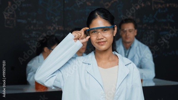 Fototapeta Highschool girl looking at camera while people doing experiment at laboratory. Young cute student pose at camera while standing blackboard with chemical theory with blurring background. Edification.