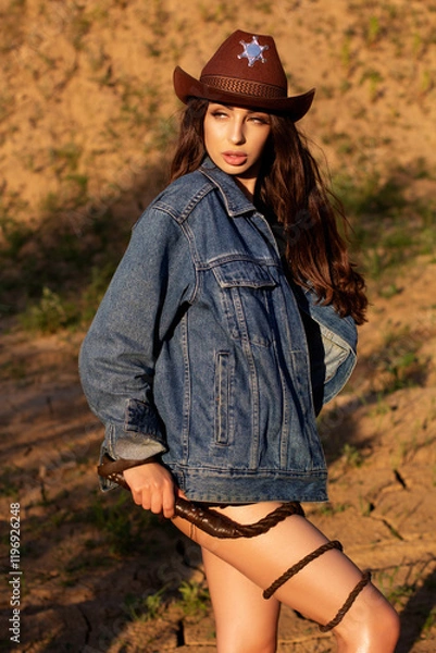 Obraz A young woman in a cowboy hat, denim jacket, and boots holding a whip and posing on cracked earth, with a dramatic western-style backdrop.