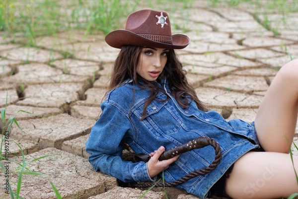Obraz A young woman in a cowboy hat, denim jacket, and boots holding a whip and posing on cracked earth, with a dramatic western-style backdrop.