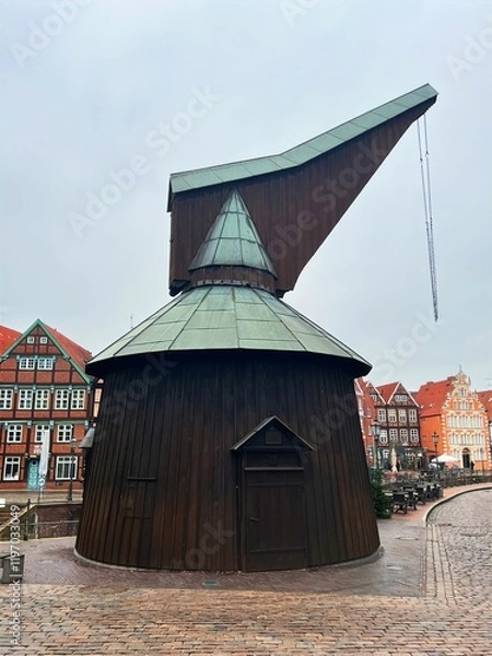 Fototapeta A historic wooden crane with a copper roof stands on a cobblestone square, surrounded by charming half-timbered buildings. The overcast sky adds a nostalgic atmosphere to the scene.