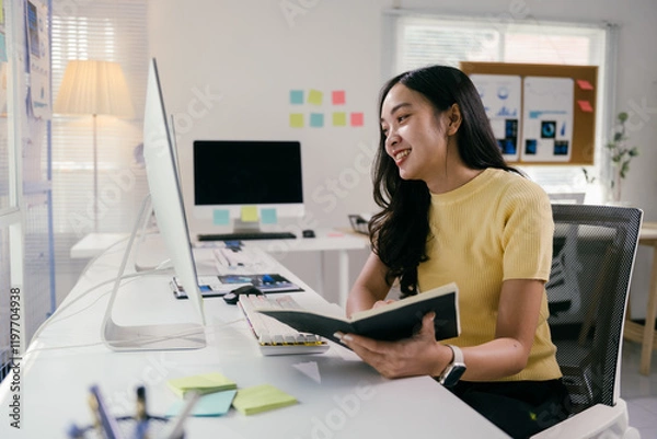 Fototapeta Young professional is smiling while working at her desk, she is holding a notebook and looking at her computer monitor