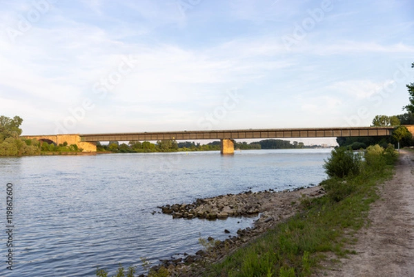 Obraz A wide view of a bridge spanning across a calm river, surrounded by lush green vegetation on both sides.