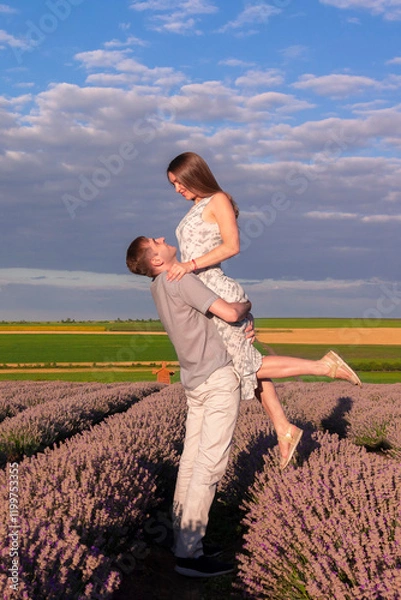 Fototapeta Joyful couple shares a romantic moment in a lavender field at sunset. The man is lifting the woman in the air as they smile warmly to each other