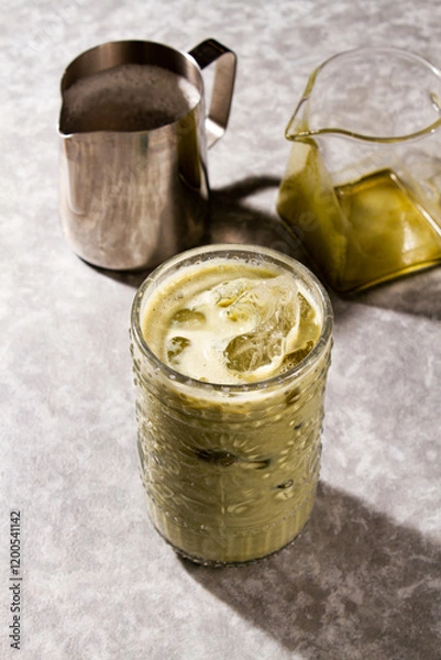 Fototapeta Iced Matcha Latte served into cold glass with ice, an empty glass jar, an a milk silver jar over a grey background