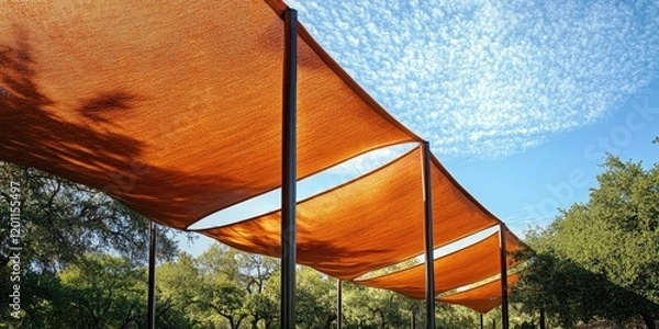 Obraz Orange shade structure positioned diagonally against a bright blue sky with white clouds, surrounded by lush green trees creating a vibrant contrast.
