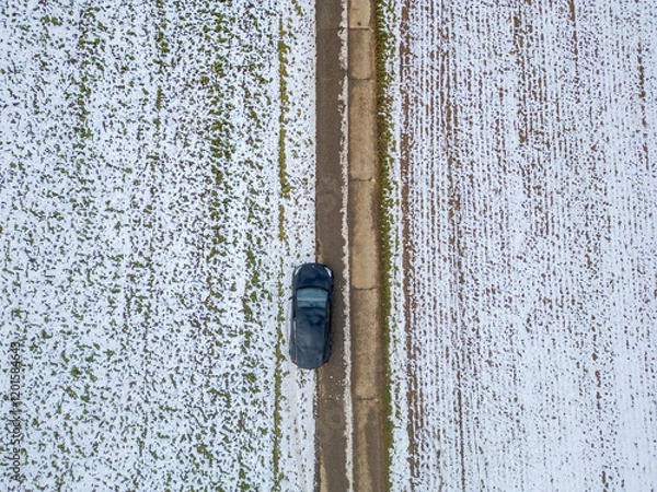 Fototapeta This aerial image captures a car navigating along a snowcovered road, which is flanked by open fields