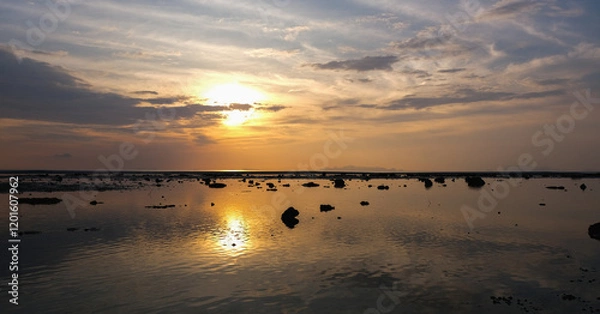 Fototapeta A serene sunset over a calm, reflective sea with scattered rocks and ripples on the water.