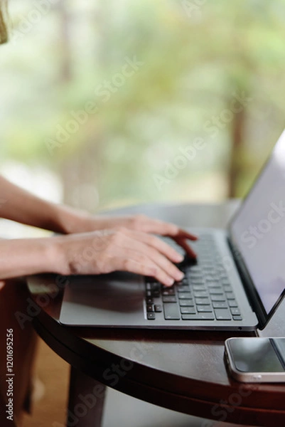 Fototapeta A young woman typing on a laptop at a wooden table in a serene outdoor setting, showcasing a relaxed atmosphere and focus on productivity in nature