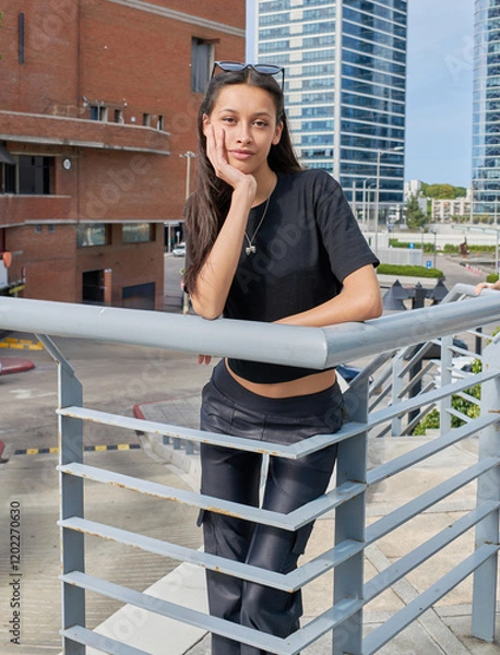 Fototapeta A young woman with dark hair and glasses on her head poses leaning on a railing, one hand on her face. She is wearing a black T-shirt and dark pants. In the background, modern urban style buildings