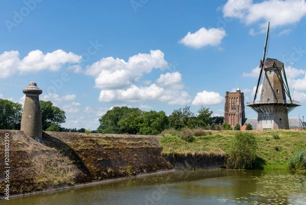 Fototapeta Fortified walls of historic town Woudrichem, the Netherlands with flour mill and characteristic square tower of Sint Martinus church against a white clouded blue sky