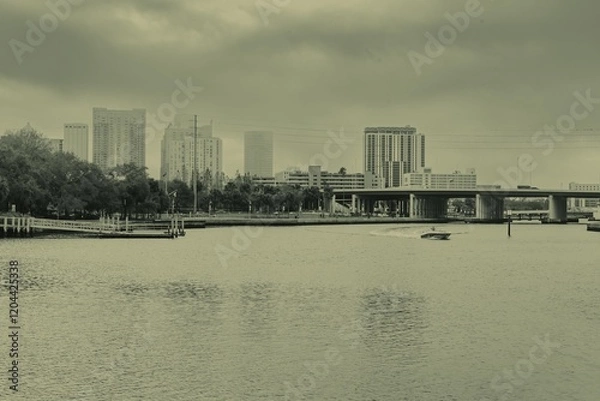 Fototapeta a boat glides up the Tampa Bay Riverwalk under a dramatic, cloudy sky, offering a tranquil view of the city’s vibrant waterfront. The calm waters reflect the changing clouds above, creating a serene a