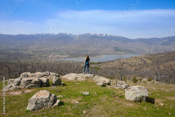 Fototapeta Woman standing with her back on a rock at the top of a mountain looking at the Lozoya Valley reservoir, with Pinilla village and the mountains of the Guadarrama National Park, in Madrid, Spain