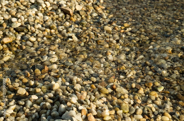 Fototapeta Pebbles and stones beneath the shallow surface of crystal-clear water. The sunlit scene creates shimmering reflections highlighting the textures and colors of the stones.