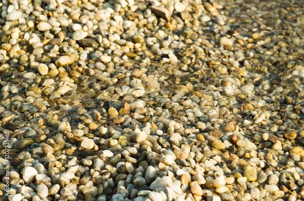 Fototapeta Pebbles and stones beneath the shallow surface of crystal-clear water. The sunlit scene creates shimmering reflections highlighting the textures and colors of the stones.