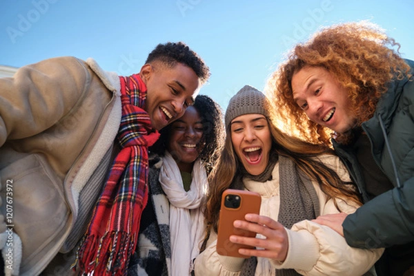 Obraz Four young cheerful multi ethnic people having fun together outdoors in winter, looking at smartphone screen