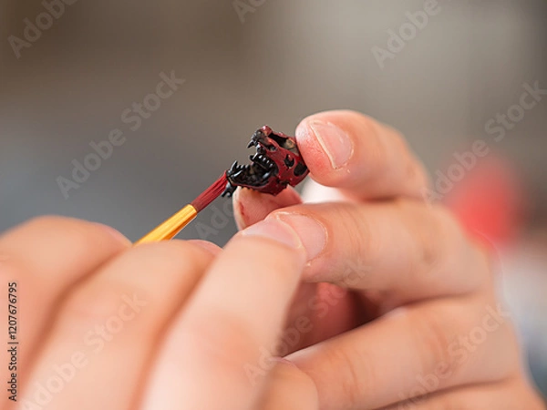Fototapeta Close-up photograph of painting a part of a wargaming model with red paint. The image focuses on the brush, the model's surface, and the fine details being applied.
