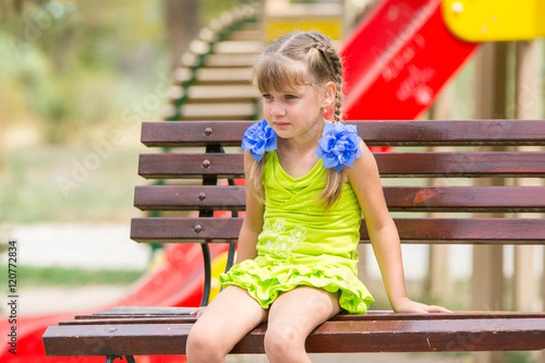 Fototapeta Portrait of upset five year old girl who is sitting on the bench on the background of the playground