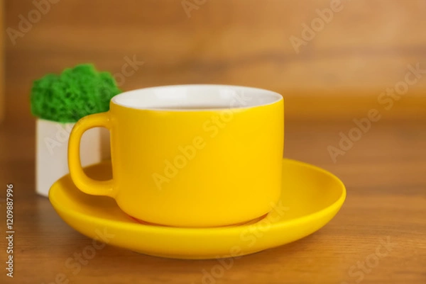 Fototapeta A yellow mug with a saucer, in which a hot drink is poured, stands on a wooden background, behind it you can see a plant in a pot view from the side. The concept of tea drinking, tea traditions. High