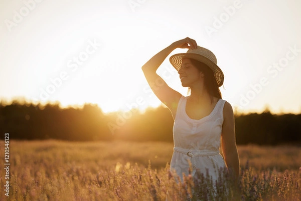 Obraz Woman in a lavender field with the sunset in the background. Young woman in white dress and straw hat, smiles and enjoys the end of the day calm and serene.